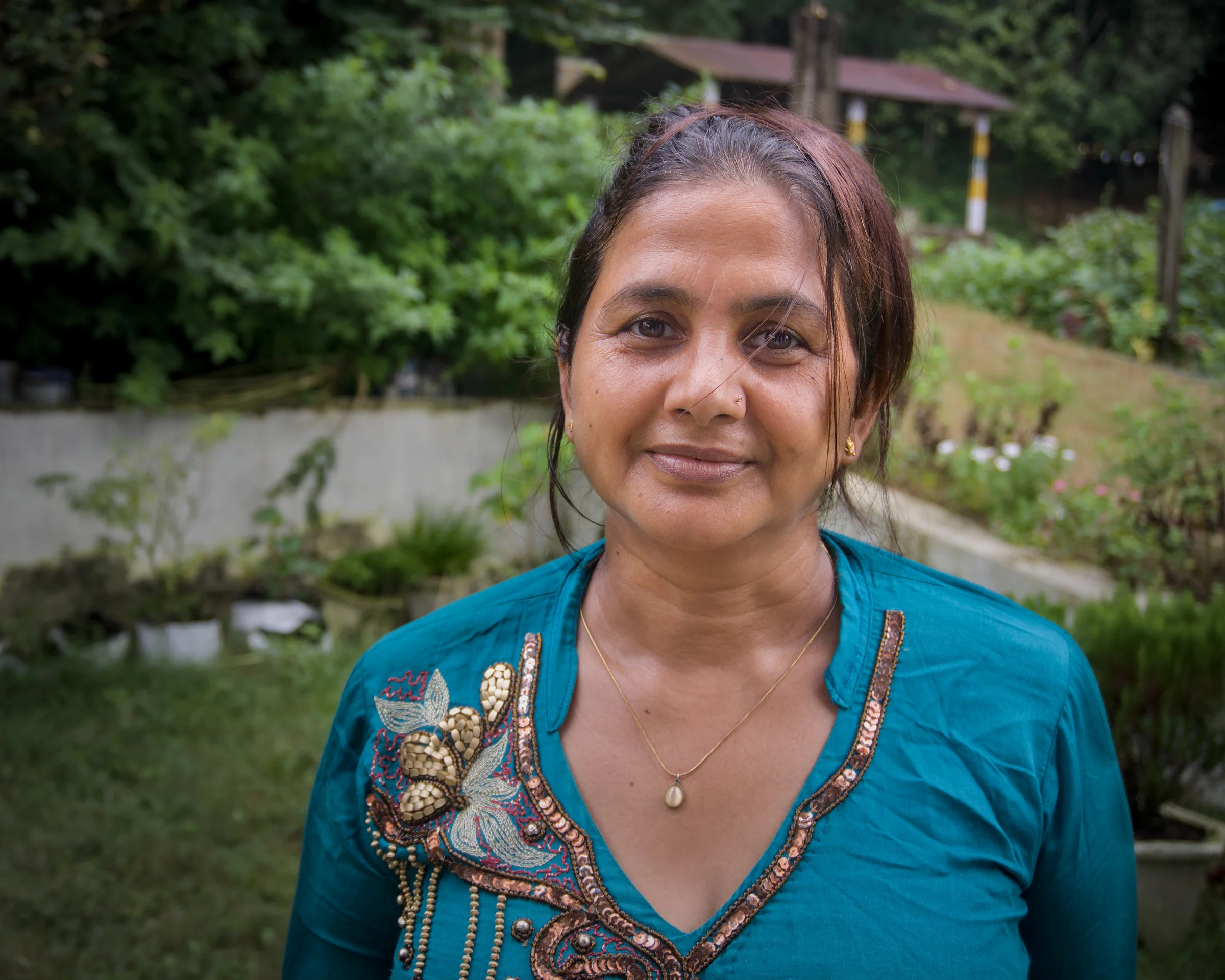 A woman in a teal embroidered kurta standing in a garden, looking at the camera, with vegetable beds and trees behind her.