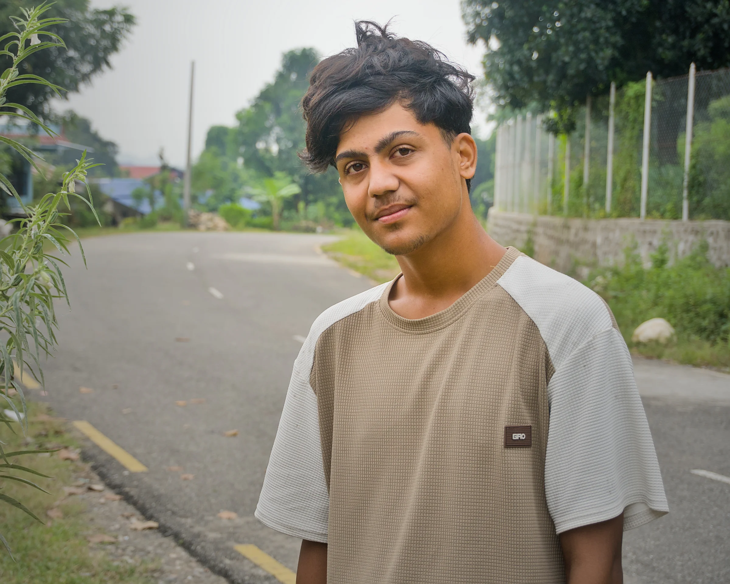 A young man in a two-tone beige t-shirt standing on an empty village road, looking at the camera, the road and houses falling away out of focus behind him.